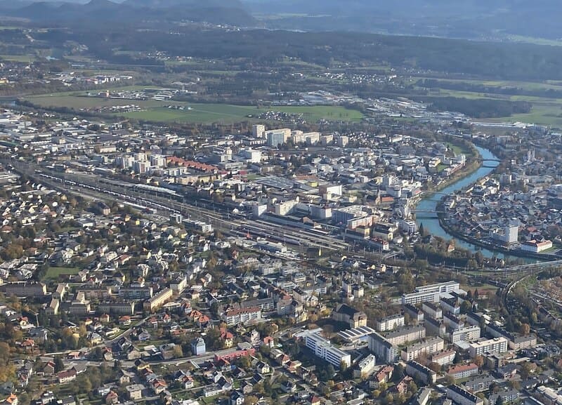 Luftaufnahme von Villach mit dicht bebautem Stadtgebiet, Flusslauf der Drau und Bahnanlagen.